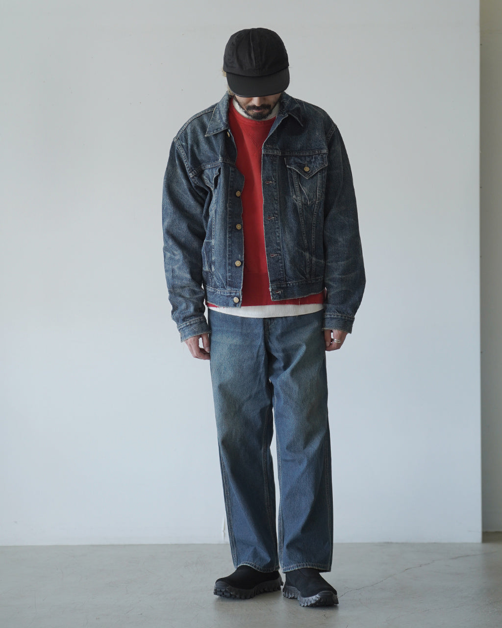 男性が denimジャケットと赤のニットを重ね着し、帽子をかぶって立つカジュアルコーディネートの全身写真