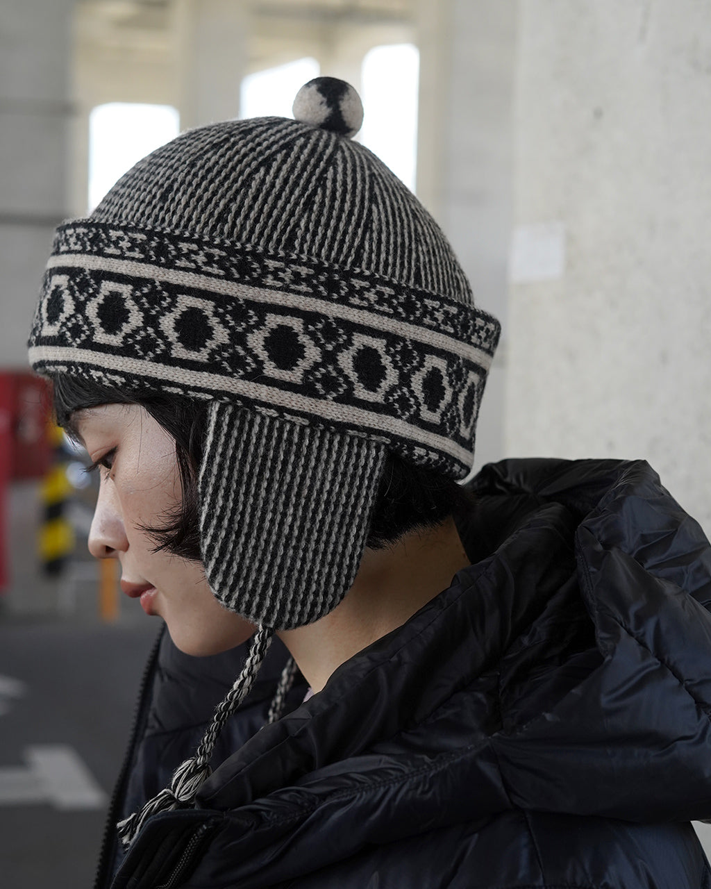 黒と白の柄編みで耳当て付きのニットキャップの着用画像。つるりとしたボンボンが頭頂部に乗り、外観はモノトーン系のデザインです。