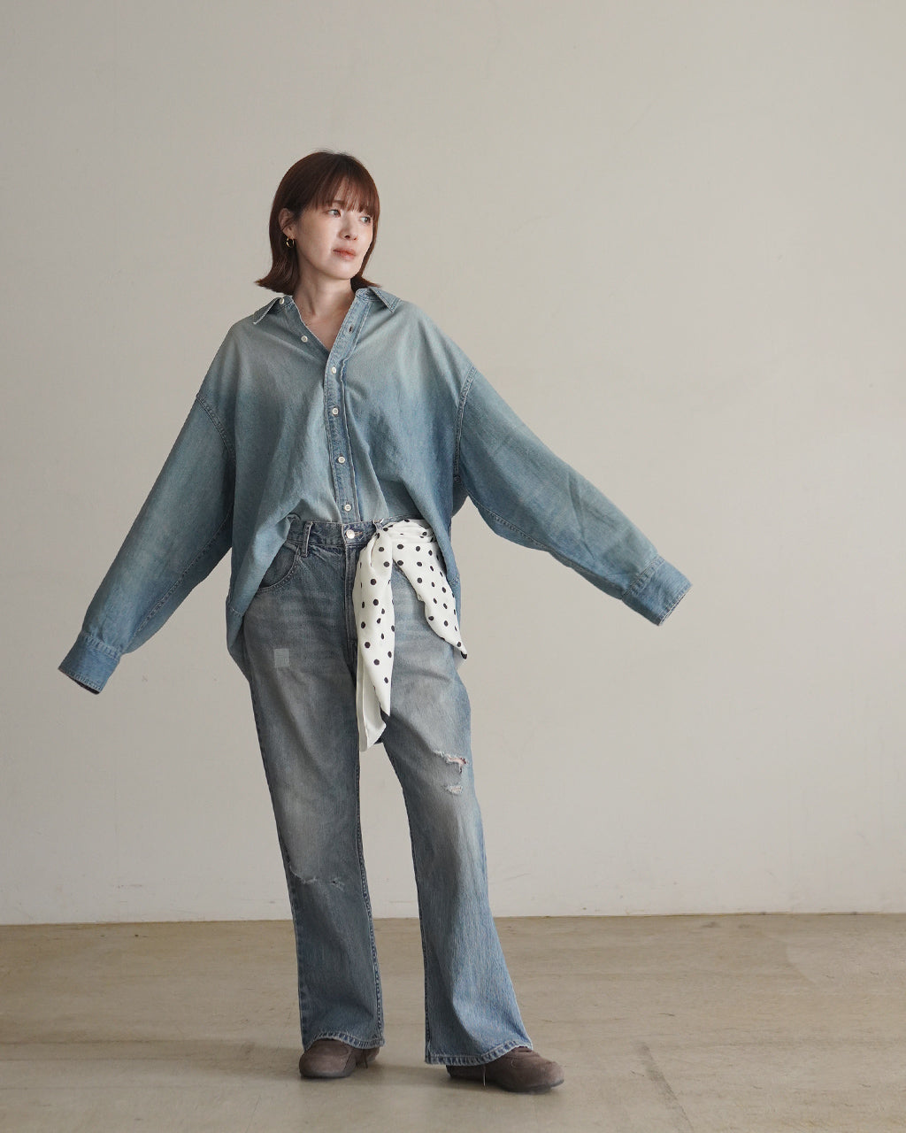 ライトブルーのOVER SIZE DENIM SHIRTを着たモデルがリラックスした雰囲気で横斜めに立つ様子。襟元が広めで前ボタン開閉、ゆとりあるシルエットが特徴的。