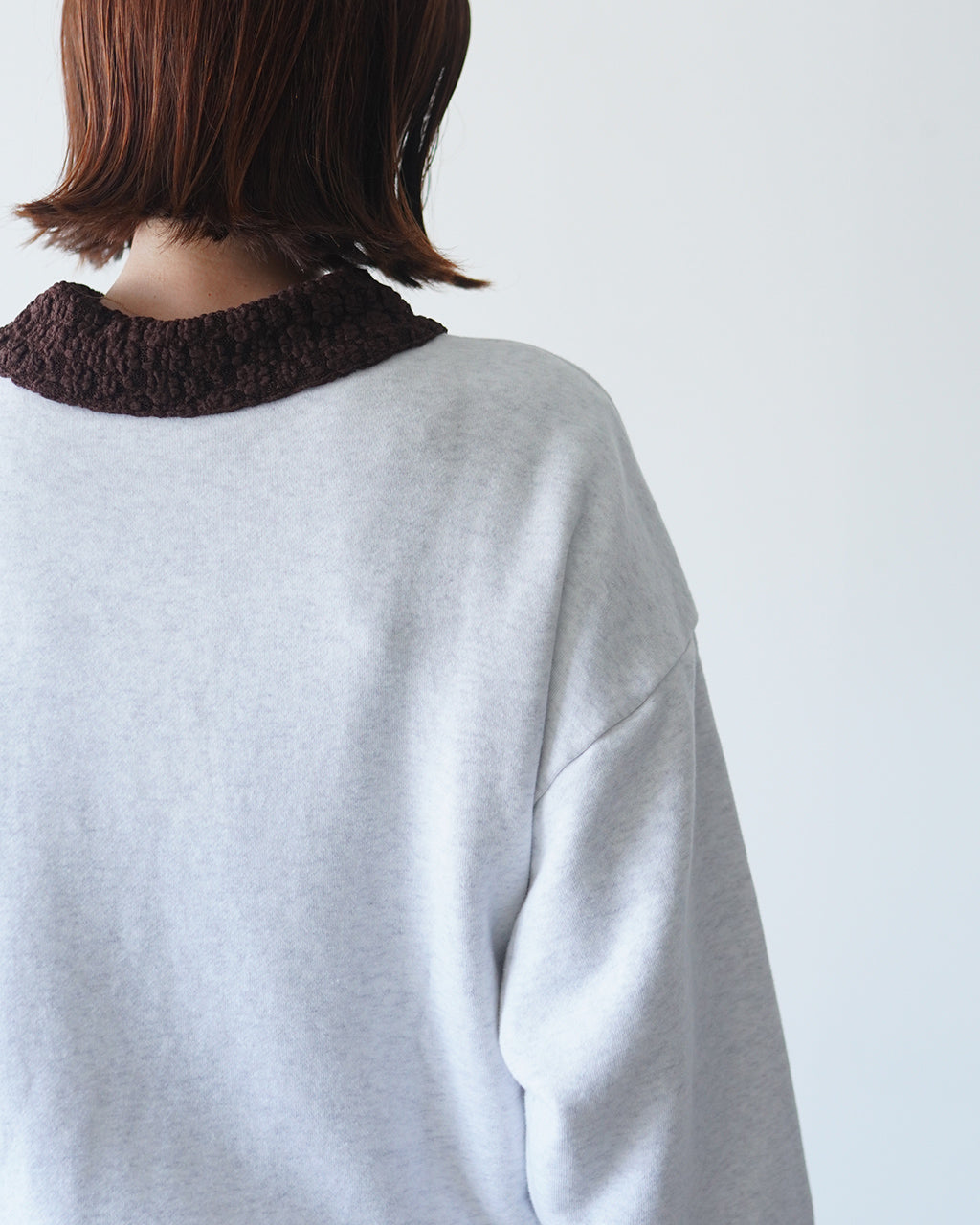シンゾーンのTHE SHELTIE SWEATSHIRT、背面寄りの画像。淡いグレーの生地にブラウンの襟リブが見え、後ろ姿が分かる。着丈は長めでゆとりのあるシルエットが印象的。