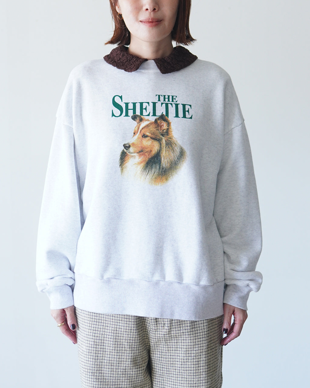 白いスウェットシャツに THE SHINZONE のロゴとシェットランド犬のプリント。首元に茶色のカラー風デザインが映えるカジュアルな着用イメージ。