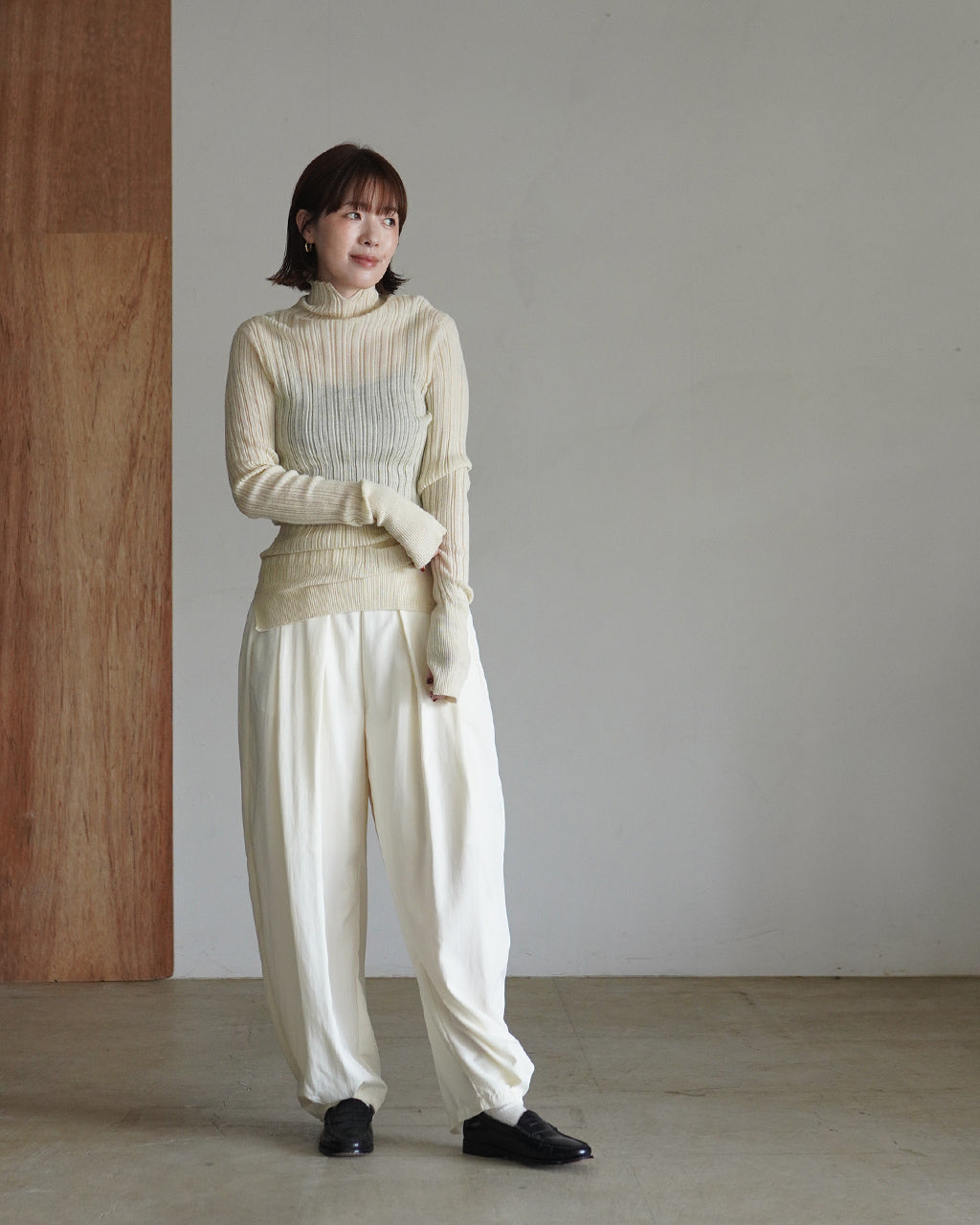淡いクリーム色のハイネックニットを着たモデルが前向きに立ち、リブ編みの表情と袖口の長めのデザインがわかる正面少し斜めのシルエット