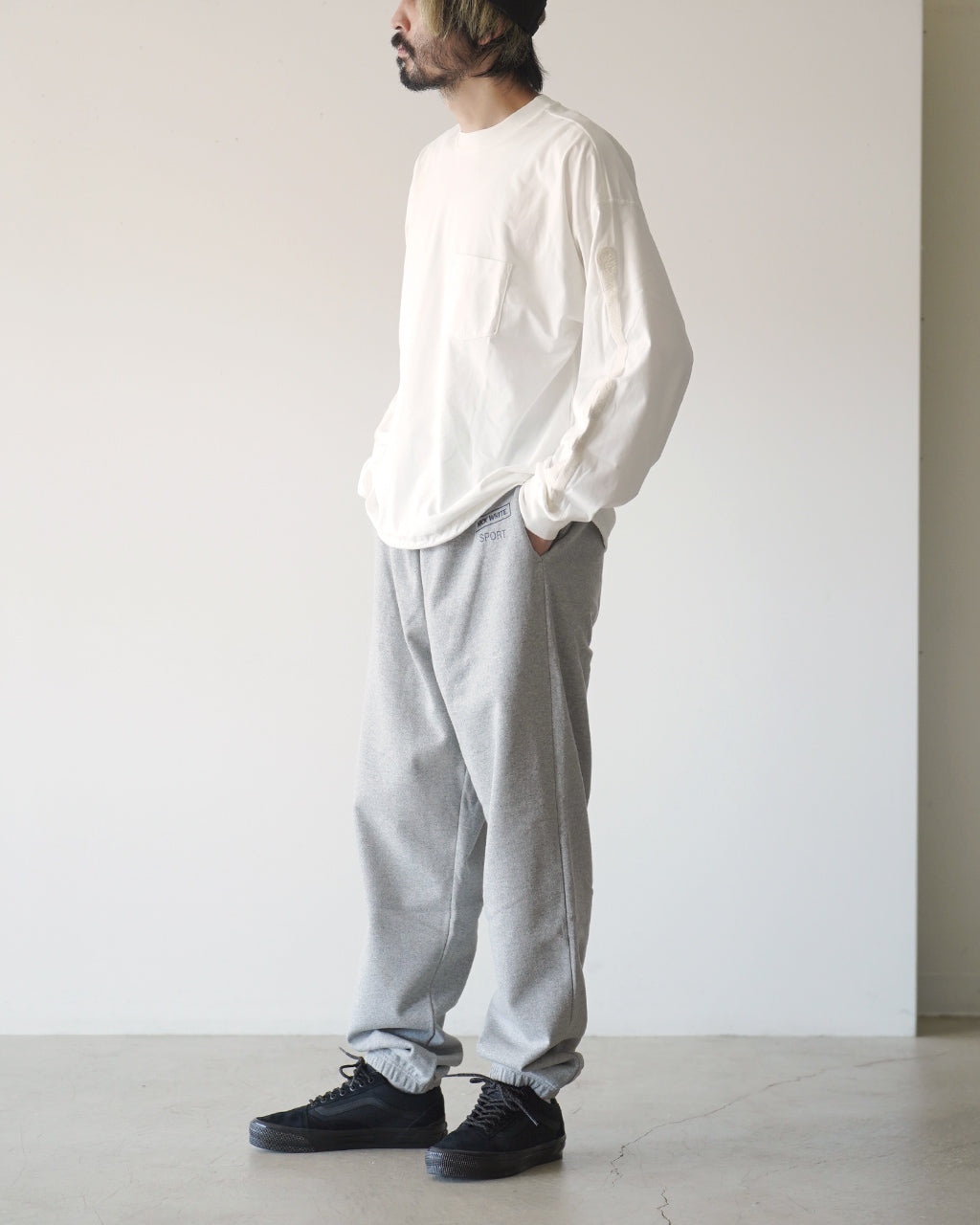 白い長袖Tシャツを着た男性が横向きに立ち、ポケット付きの前身頃が見えるカット。シンプルなリラックスシルエットで、袖口は自然に絞られたデザインが感じられる。