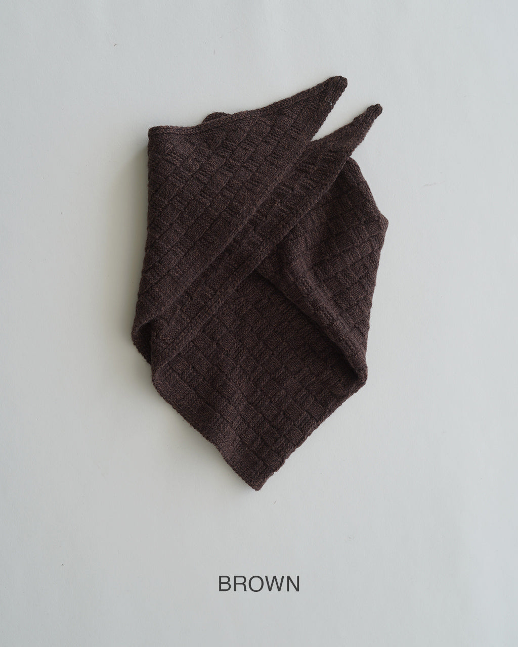 ブラウン系の BLOCK PATTERN SCARF が折りたたまれた状態。特徴的な格子柄のニット生地とコンパクトな三角形フォルムが見える。柔らかな質感と厚みを感じさせる糸組みが表現されたマフラー